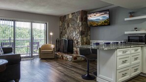 Una sala moderna con una pared de acento de piedra, una chimenea, un ventanal grande y una zona de cocina con salpicadero de piedra y armarios blancos.