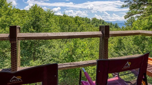 Un área de estar al aire libre con valla de madera y sillas, con vista a un bosque frondoso y un cielo despejado.