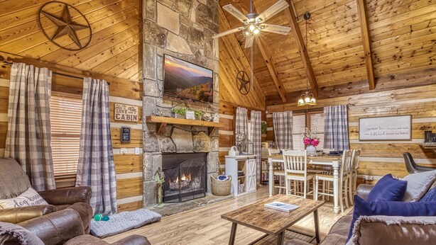 Un intérieur de chalet chaleureux avec une cheminée en pierre, des canapés en cuir, un coin repas et un ventilateur de plafond.