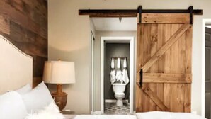 Quarto com cabeceira de madeira, cama com roupa de cama branca, mesa de cabeceira com abajur e porta de correr de madeira para um banheiro.