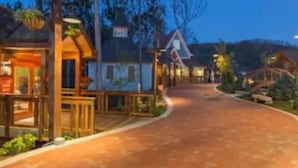 Uma trilha pavimentada conduz a um edifício de madeira com uma placa, cercado por árvores e uma paisagem acidentada ao fundo.