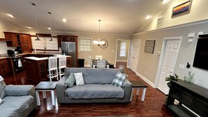 A living room with a grey sofa, a coffee table, and a television. There is a kitchen in the background with wooden cabinets and a refrigerator. - Pet Friendly Fire Pit Movie Theater Pool Sauna (Sevierville)