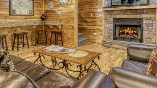 A log cabin interior with a stone fireplace, leather seating, and a television mounted above the fireplace.