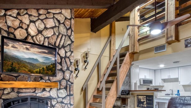 Ein Kamin aus Stein, ein Fernseher, eine Treppe, eine Küche und ein Ledersofa in einem Wohnzimmer.