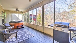 A covered deck with a hot tub, outdoor seating, a grill, and large windows offering a view of trees and a building. - Irresistible Retreat w Pool Hot Tub Arcade (Sevierville)