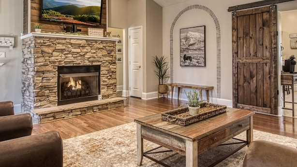 A living room with a stone fireplace, a mounted TV, a ceiling fan, and a rustic wooden door. - Irresistible Retreat w Pool Hot Tub Arcade (Sevierville)