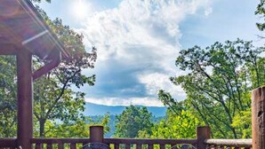 Une terrasse en bois avec une table ronde et des chaises en métal, entourée d’une balustrade en bois, d’où l’on peut admirer une zone boisée avec des montagnes à l’horizon.