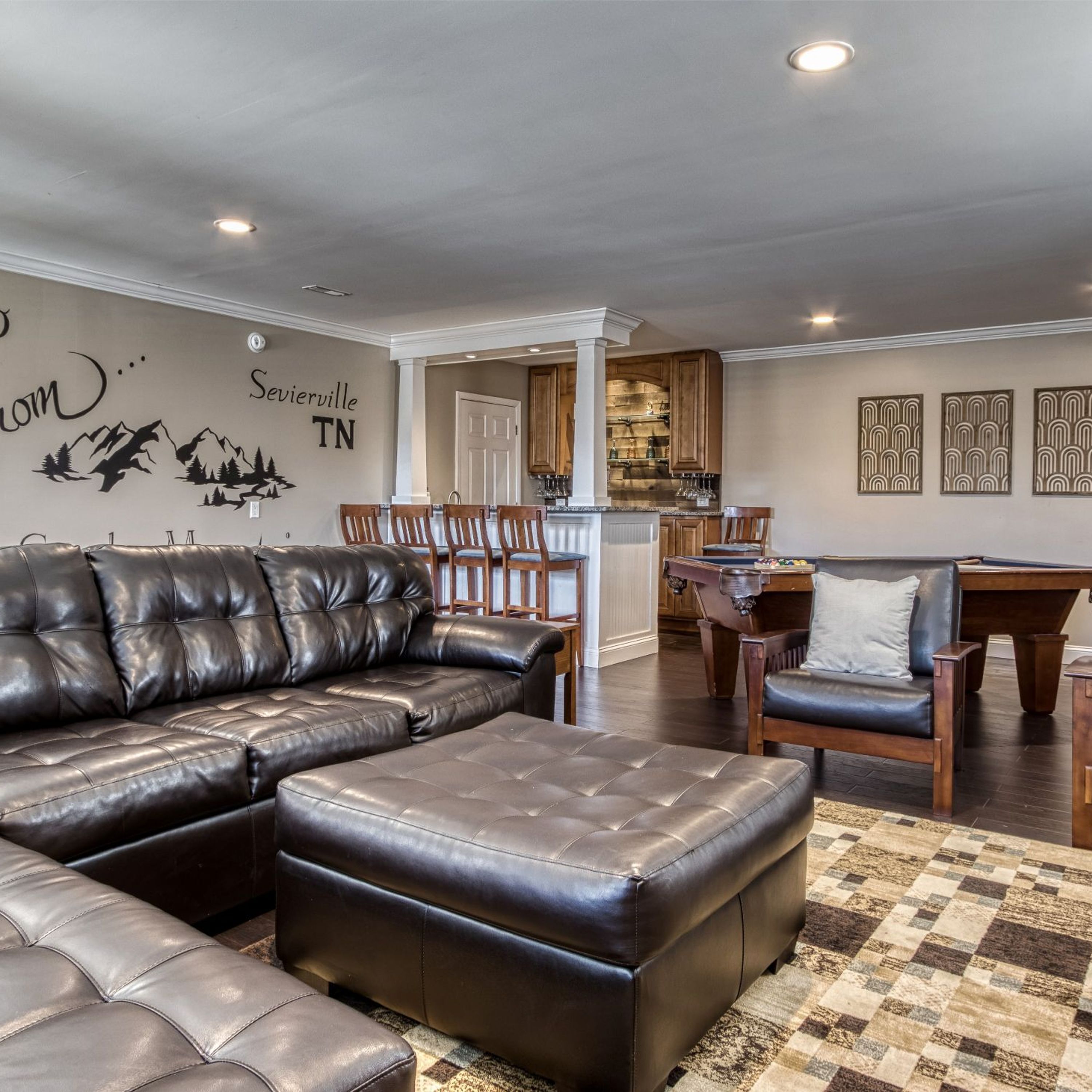 A living room with a sectional sofa, ottoman, and a pool table. There is a kitchen area with wooden cabinets and a dining table.