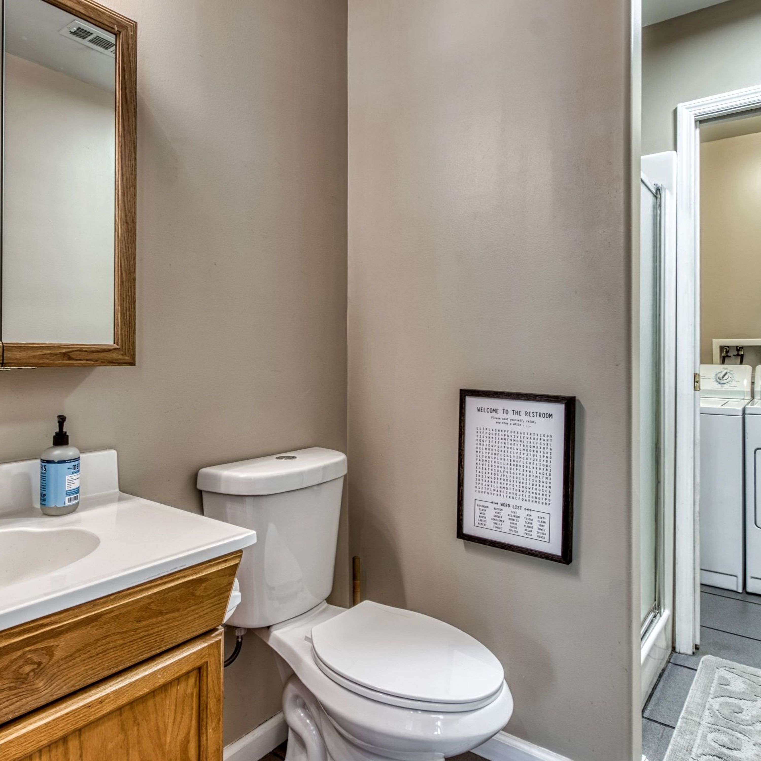 A bathroom with a toilet, a sink with a wooden vanity, a mirror, and a word list on the wall.