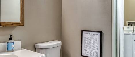 A bathroom with a toilet, a sink with a wooden vanity, a mirror, and a word list on the wall.