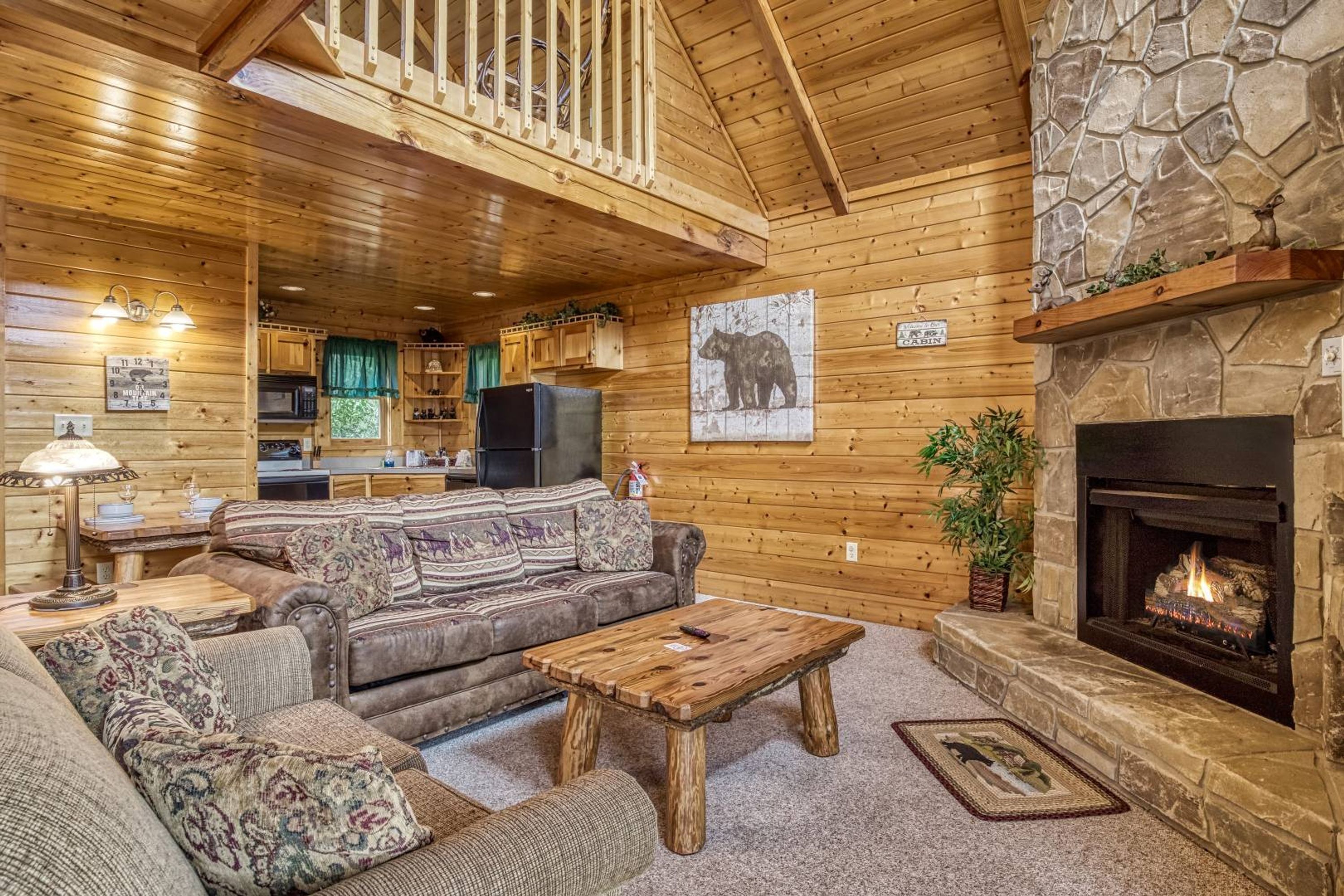 A cozy cabin interior with a stone fireplace, wooden walls, a living area with a sofa and coffee table, and a kitchen in the background.
