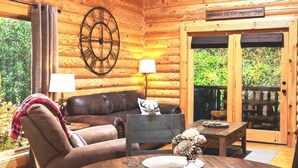 A cozy log cabin interior with a leather sofa, a round wooden table with placemats, a wall clock, and a sign reading "Welcome to the Mountains". - Cozy Upscale Cabin w Fireplace Hot Tub (Sevierville)