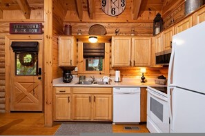 Interior de una cabaña de madera con cocina, que incluye armarios de madera, un refrigerador blanco, una estufa y un reloj en la pared.
