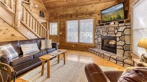 L’intérieur d’un chalet en bois, avec une cheminée en pierre, un canapé en cuir, un téléviseur et un escalier.