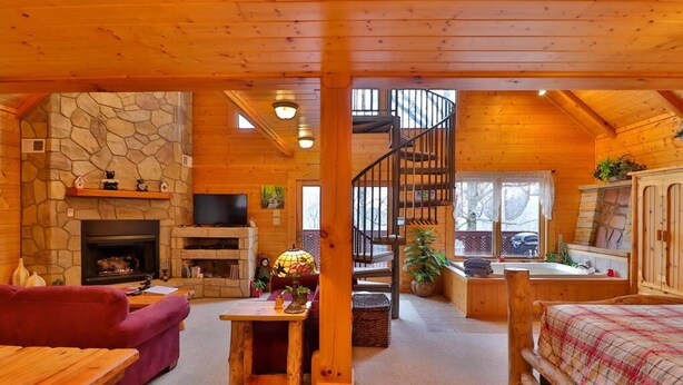 Un interior acogedor de cabaña con chimenea de piedra, mobiliario de madera, una escalera espiral y un ventanal grande.