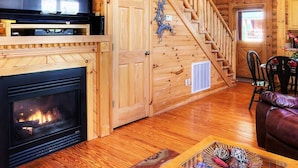 Interior de una cabaña de madera con chimenea, un televisor montado arriba, una escalera que sube a un nivel superior y un comedor con sillas.