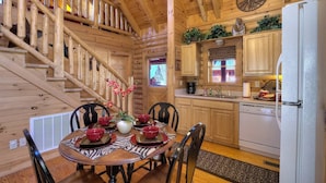Interior de una cabaña de madera con una mesa de comedor para cuatro, una cocina con electrodomésticos blancos y una escalera con barandilla de madera.