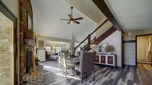A spacious living area with a dining table, chairs, and a fireplace. A ceiling fan is visible, and the room has a wooden floor. - New Luxury Remodel Fire Pit Game Room Hot Tub (Sevierville)