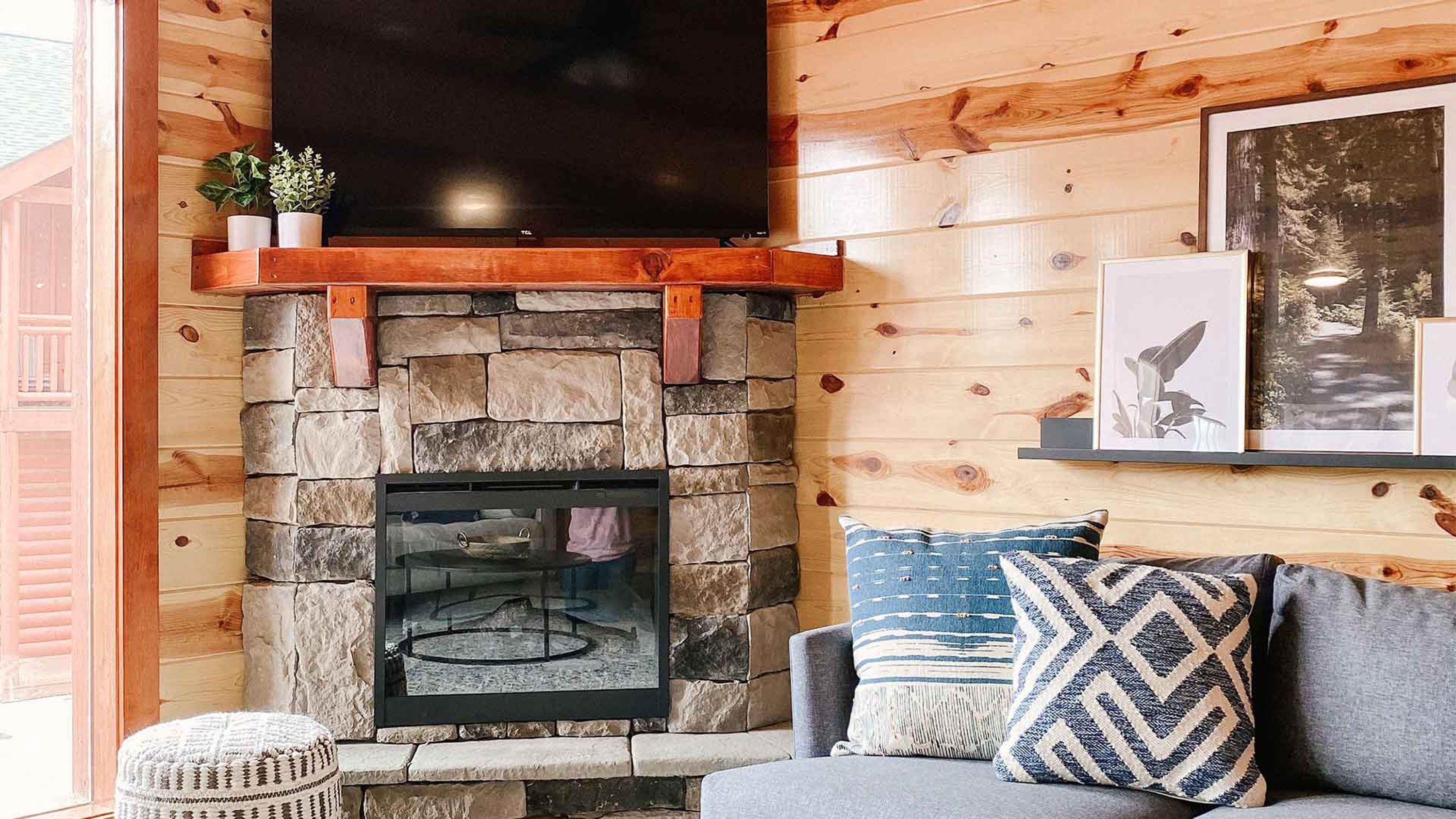 A cozy living room with a stone fireplace, a flat-screen TV mounted above it, and a comfortable seating area with cushions.