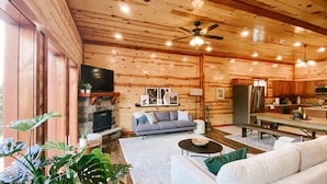 A cozy log cabin interior with a stone fireplace, a gray sofa, a dining table, and a kitchen area. - Modern Luxury w Pool Theater Hot Tub Ent Room (Sevierville)