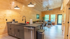 A modern kitchen with wooden walls, a central island, stainless steel appliances, and a dining area with a view. - Brand New 2 bed 2 Bath Arcade Games Hot Tub (Sevierville)
