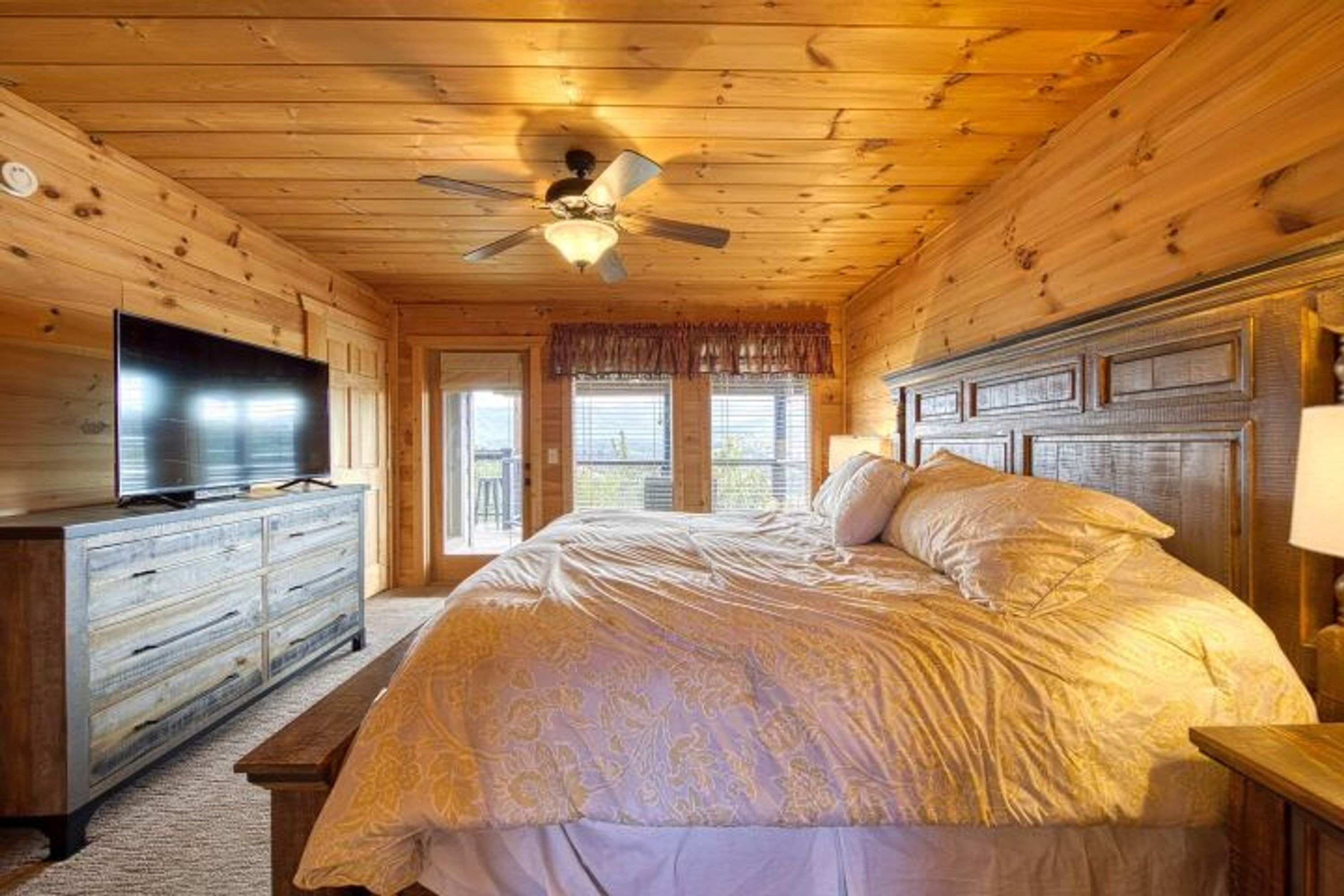 A wooden cabin bedroom with a bed, dresser, television, and a ceiling fan.