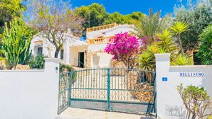 Una entrada protegida por una reja a una propiedad llamada Bellisimo, con un edificio blanco, un muro de piedra y diversas plantas, incluyendo una buganvilla rosada destacada.