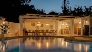 Un espace aménagé au bord de la piscine, comprenant une table et des chaises, une pergola agrémentée de guirlandes lumineuses et un coin cuisine avec un barbecue.