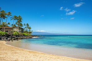 A beachfront hotel with a sandy beach, palm trees, and ocean views. - Kahana Villas E408 by Gather: Prime Location w/ Pool,Beach,Tennis & Sunset Views (Lahaina)