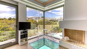 A modern living room with a television, clear chairs, a small table, and a glass pool area. - A2 -  luxury house with pool (Porto Heli)