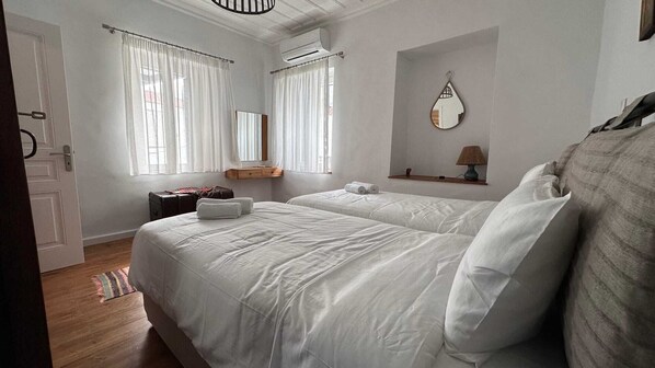 Un dormitorio con dos camas, piso de madera, una mesita con una lámpara y una ventana con cortinas.