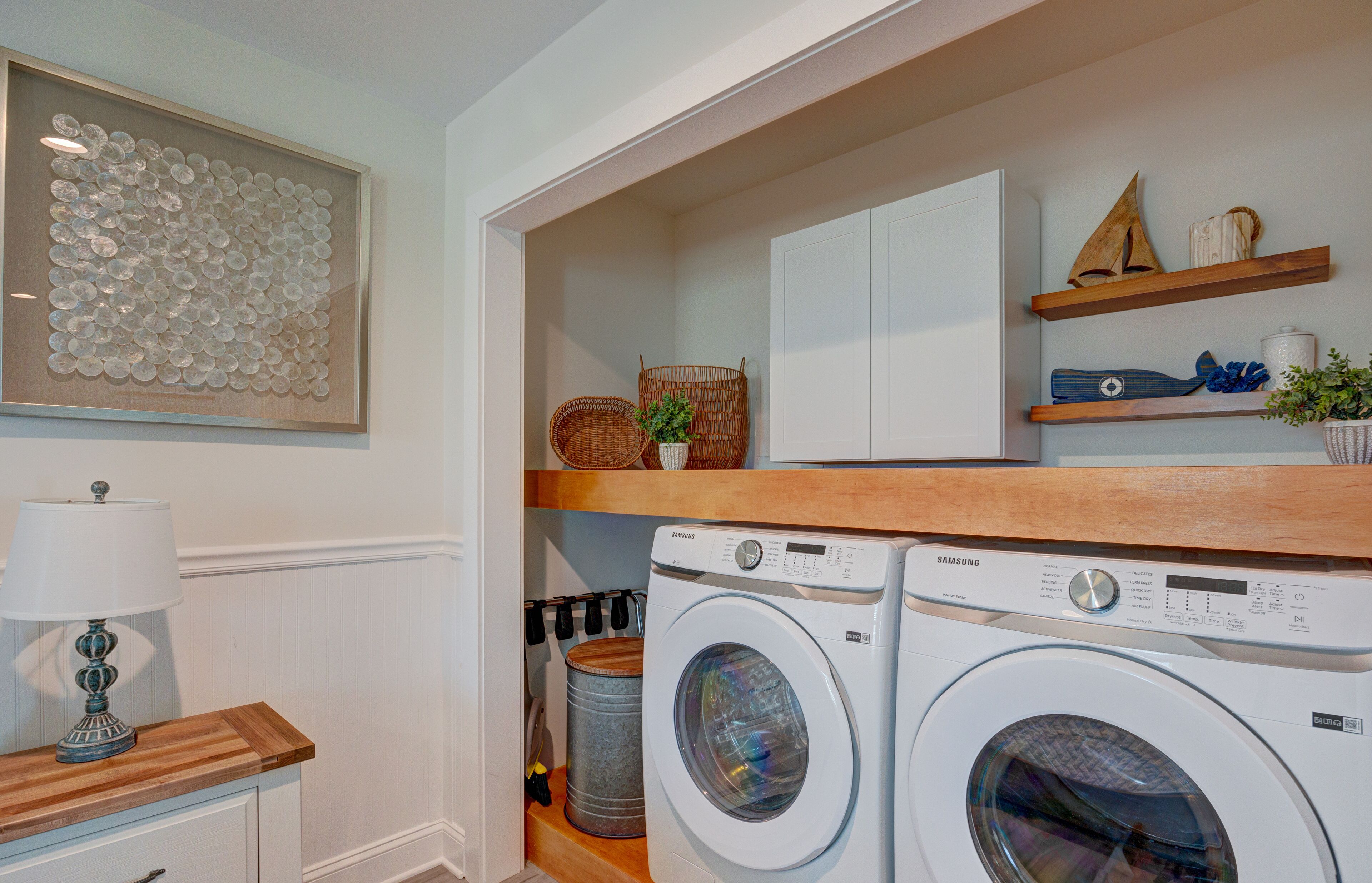 A laundry room with two white front-loading washing machines, a wooden shelf with decorative items, a potted plant, and a framed artwork on the wall.
