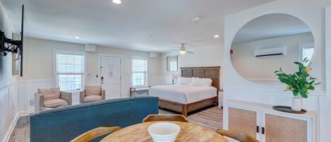 A modern hotel room with a large bed, a seating area with a blue sofa, a dining table with chairs, and a large mirror.