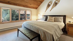 A bedroom with a large bed, a bench, two wall-mounted decorative items, and a window with a view of trees. - Charming and Upgraded 3 Bedroom Mountain Townhome by Gorecreek Rentals (Vail)