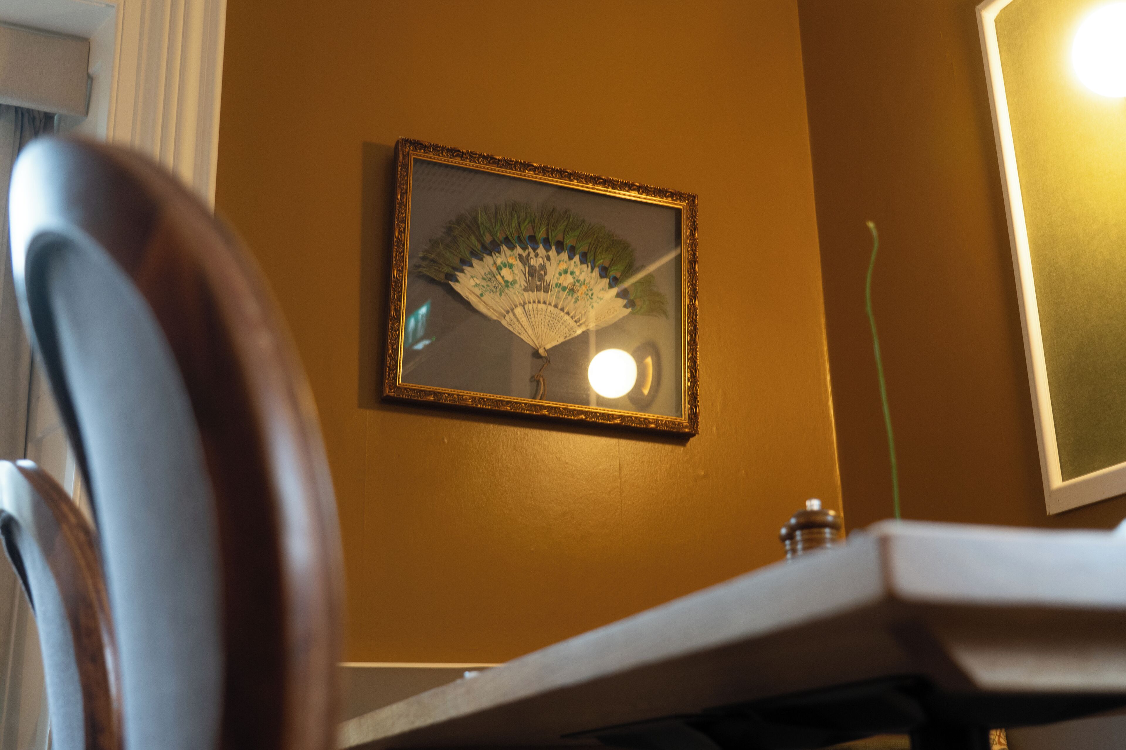 A framed fan on a wall, a chair, and a table with a lamp.