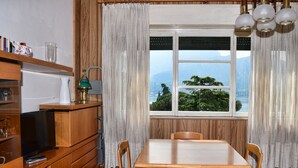 A dining area with a table and chairs, a television on a wooden cabinet, a lamp, and a window with a view of trees and mountains. - Coral Blue by Wonderful Italy (Bellagio)