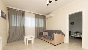 A living room with a beige sofa, a small white table, and a bed with patterned bedding in the background. - Cabeceo in Jerez de la Frontera (Jerez de la Frontera)