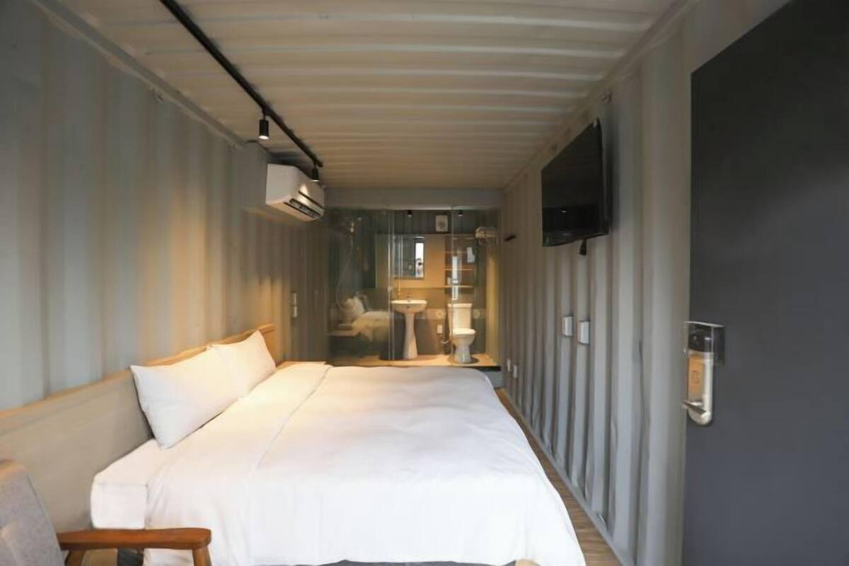 A narrow, elongated room with a bed, a small table, and a television mounted on the wall.