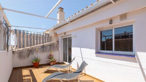 En solbeskinnet terrasse med to liggestole, en lille plantekasse og et vindue med udsigt til et hegn.