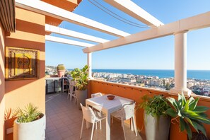 Ein Balkon mit weißem Tisch und Stühlen, Blumentöpfen und Blick auf das Meer und die Stadt.