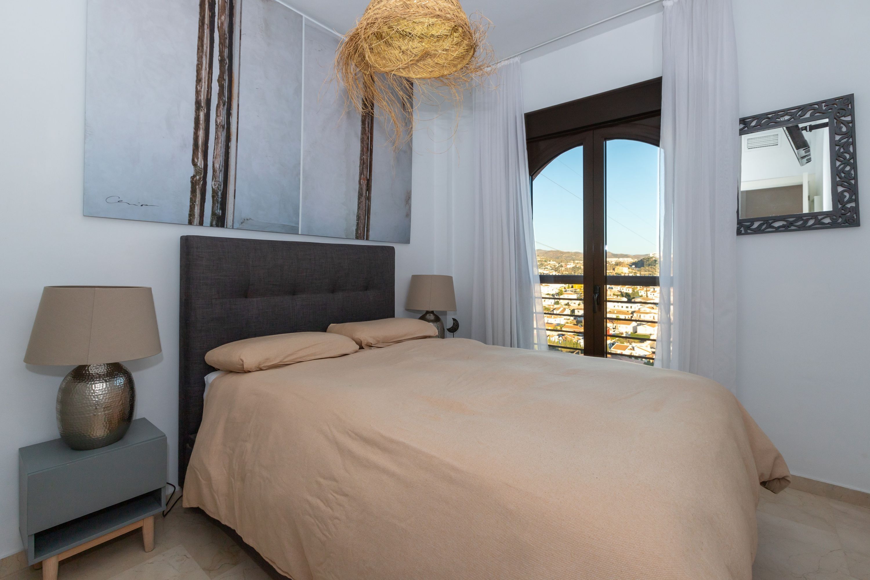 A bedroom with a bed, two bedside tables with lamps, a large window with a view, and a wall-mounted mirror.