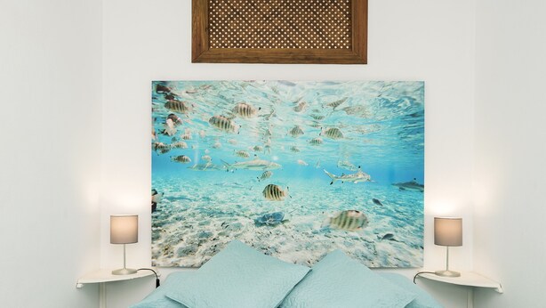 Een slaapkamer met een groot schilderij met een zeethema, een bed met lichtblauwe beddengoed en twee bedlampjes.