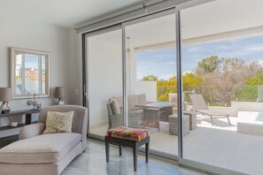 A modern living room with a sofa, a small table with a colorful cushion, and a sliding glass door leading to an outdoor patio with a pool and lounge chairs. - Artola in Marbella With 4 Bedrooms and 3 Bathrooms (Marbella)