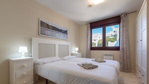 Et soveværelse med to senge, et natbord med lampe, en radiator og et vindue med gardiner.