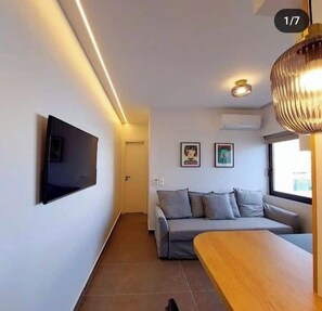 Ein modernes Wohnzimmer mit einem an der Wand befestigten Flachbildfernseher, einem grauen Sofa, einem Holztisch und einer Pendelleuchte.