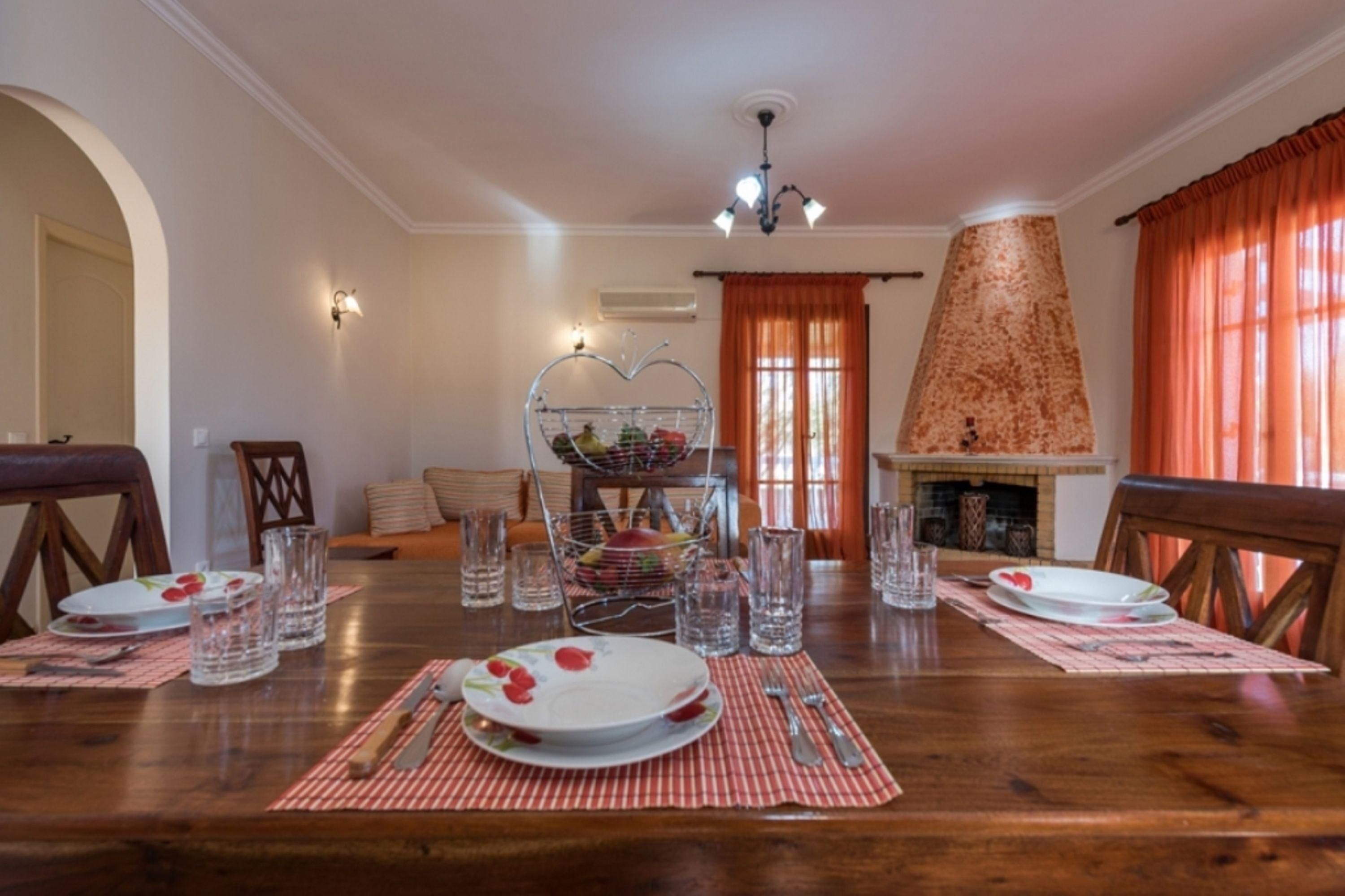 A dining area with a wooden table set for four, surrounded by chairs. There is a fireplace with a decorative stone mantle, and a wall-mounted light fixture above the table.