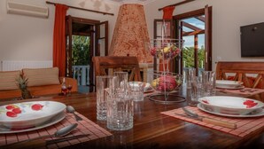 A dining area with a wooden table set for a meal, a fruit basket, and a view of greenery outside. - Villa Rose With Private Pool (Zakynthos)