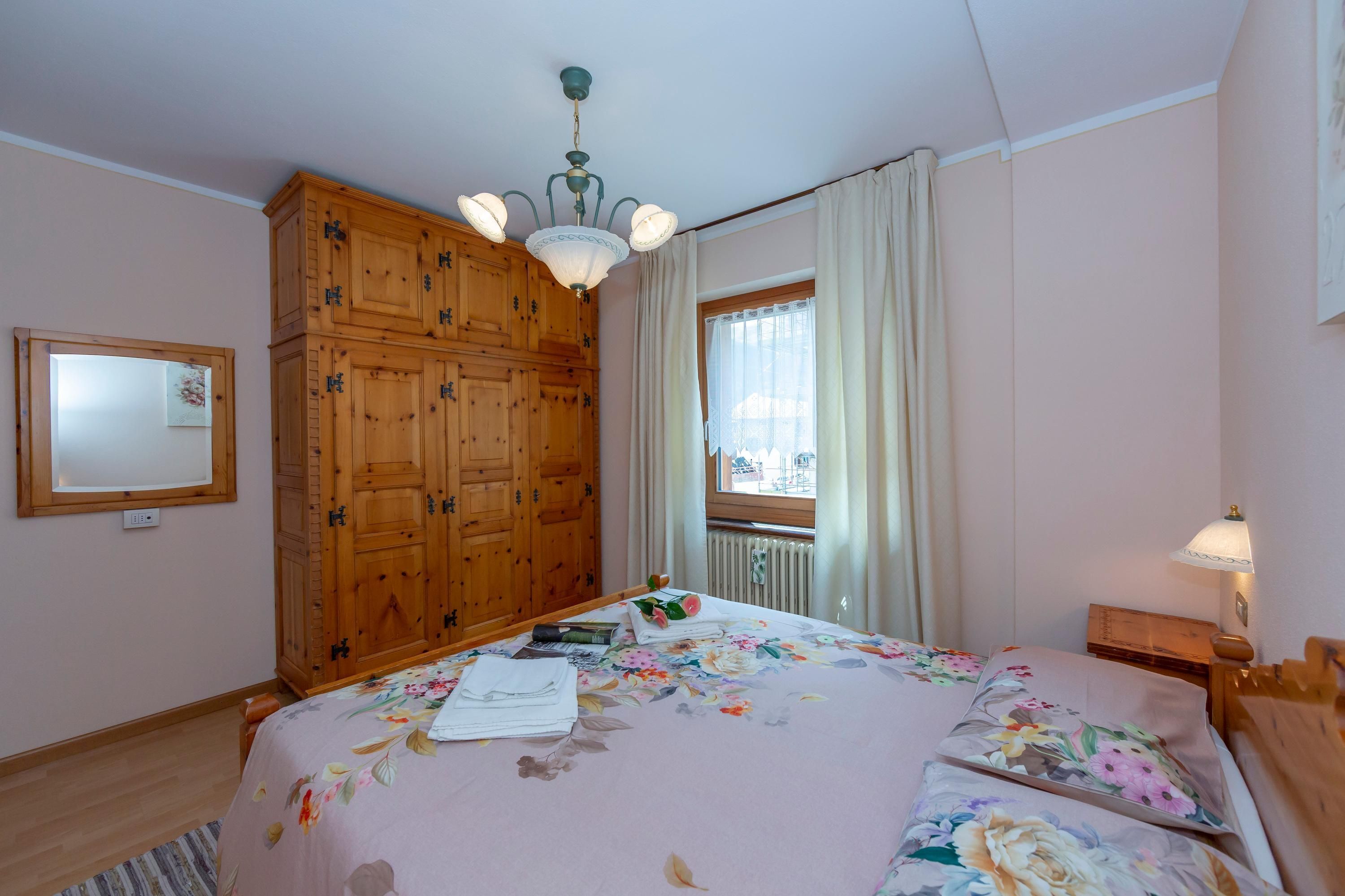 Ein Schlafzimmer mit großem Holzschrank, einem Bett mit Blumenbettwäsche, einem Fenster mit Vorhängen und einer Deckenleuchte.
