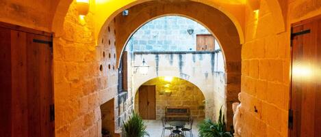 An arched corridor with stone walls, a wooden door on the left, and a bench with chairs in the center.