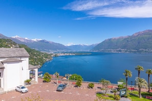 A coastal view with a building, parked cars, and a clear blue lake. - Red View Apartment in Ronco Sopra Ascona Ascona (Ronco sopra Ascona)